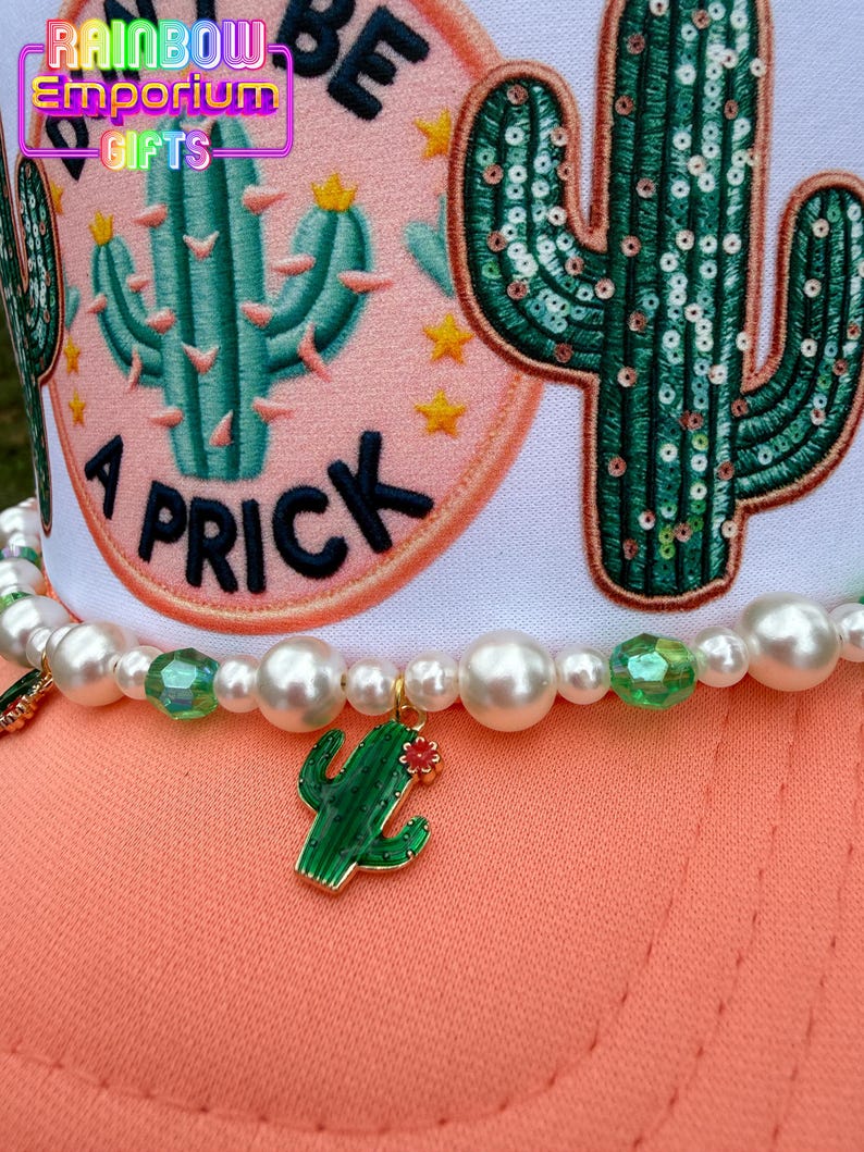 May include: A white and peach-colored hat with a beaded necklace. The hat features a cactus design and the text "A Prick". The necklace has a green cactus charm and pearl beads.
