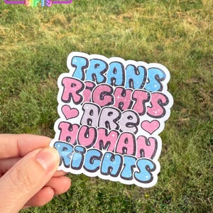May include: A sticker with the text "Trans Rights Are Human Rights" in a colorful, bubbly font. The sticker is pink, blue, and white with glitter accents.