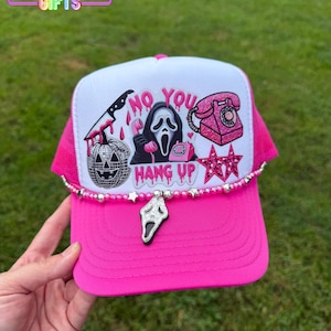 May include: A hot pink trucker hat with a white front panel featuring a Scream mask, a bloody knife, and the words "NO YOU HANG UP." The hat has a silver charm and a pink mesh back.