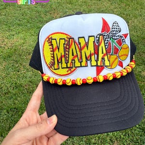 May include: Black and white baseball cap with the word "MAMA" in yellow, with a softball design. The cap has a red and black checkered bow and yellow and red beads along the brim. The text "Rainbow Emporium Gifts" is in the upper left corner.