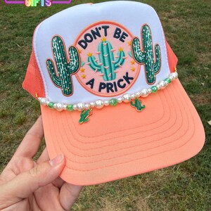 May include: A coral and white trucker hat with a "Don't Be a Prick" cactus design. The hat features a beaded trim with cactus charms and the text "Rainbow Emporium Gifts".