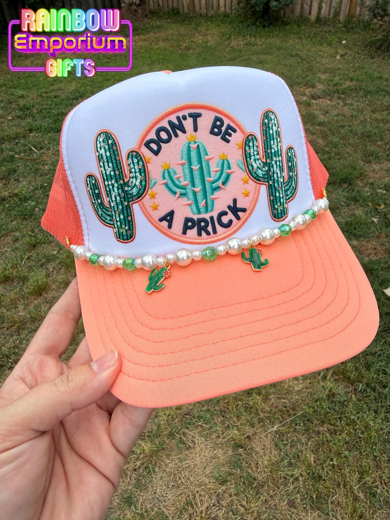 May include: A coral and white trucker hat with a "Don't Be a Prick" cactus design. The hat features a beaded trim with cactus charms and the text "Rainbow Emporium Gifts".