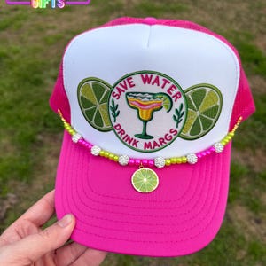 May include: Pink and white trucker hat with a lime green and white embroidered design that says "Save Water Drink Margs". The hat has a beaded trim with a lime green charm.