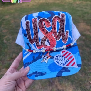 May include: A blue camouflage trucker hat with the word "USA" in red sequins. The hat features a white mesh back, a beaded accent, and patriotic charms, including a star and a heart.