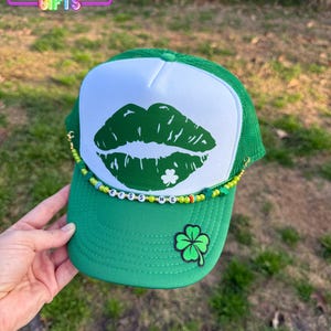 May include: Green trucker hat with a white mesh back. The front of the hat features a green lip print with a shamrock in the center. The hat has a green beaded band with the words "Kiss Me" in white beads.