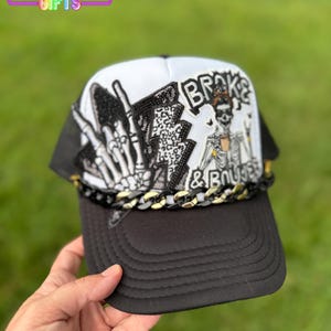May include: Black and white trucker hat with a skeleton graphic and the words "Broke & Boujee". The hat features a black brim, a white front panel, and a black mesh back. A gold chain detail accents the brim. The hat is from Rainbow Emporium Gifts.
