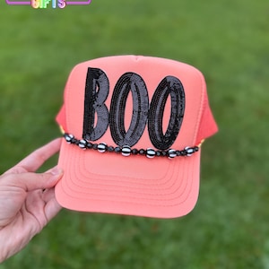 May include: Coral-colored trucker hat with the word "BOO" spelled out in black sequins. The hat features a beaded band with black and white beads. The hat is held up by a hand. The text "RAINBOW Emporium GIFTS" is in the background.