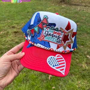 May include: A red, white, and blue trucker hat with patriotic embellishments. The hat features a red brim, a white crown, and a blue mesh back. Embellishments include a heart-shaped American flag, stars, and the text "She loves Jesus & America too."