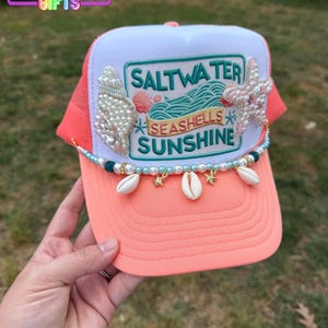May include: A coral and white trucker hat with a mesh back. The hat features a patch with the words "Saltwater, Seashells, Sunshine." Embellishments include pearls, seashells, and starfish. The hat is decorated with a beaded strand and charms.