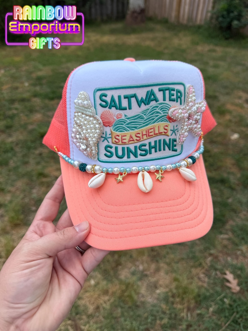May include: A coral and white trucker hat with a mesh back. The hat features a patch with the words "Saltwater, Seashells, Sunshine." Embellishments include pearls, seashells, and starfish. The hat is decorated with a beaded strand and charms.
