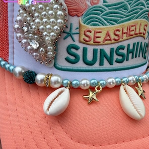 May include: A coral-colored baseball cap with a white front panel featuring embroidered text and a seashell design. The cap is adorned with a pearl and rhinestone pendant, a beaded strand with seashells and gold starfish charms. The text on the cap reads "Seashells Sunshine".