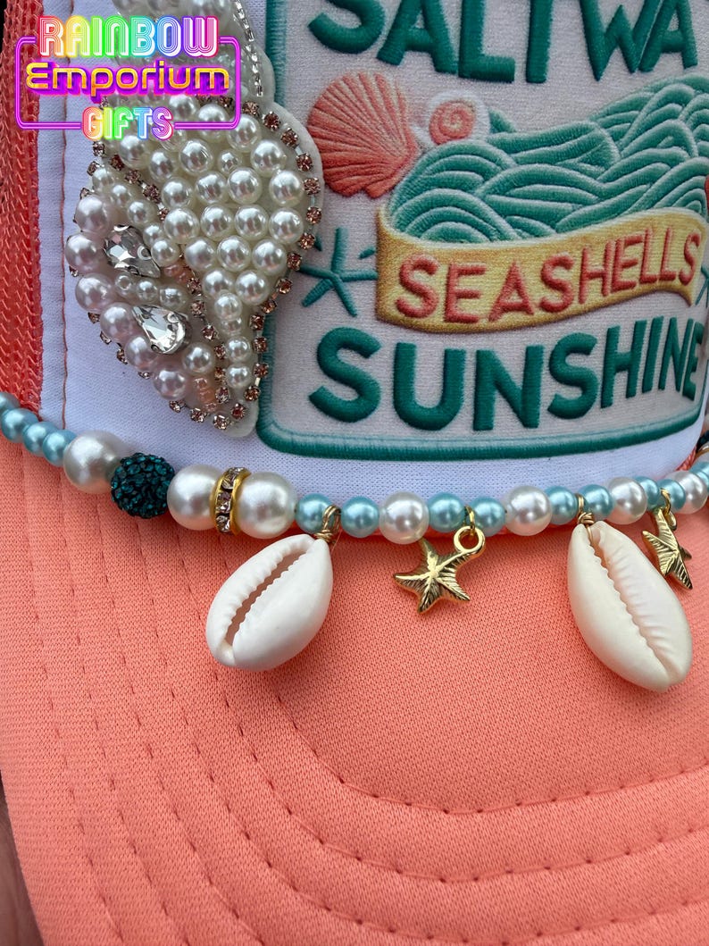 May include: A coral-colored baseball cap with a white front panel featuring embroidered text and a seashell design. The cap is adorned with a pearl and rhinestone pendant, a beaded strand with seashells and gold starfish charms. The text on the cap reads "Seashells Sunshine".