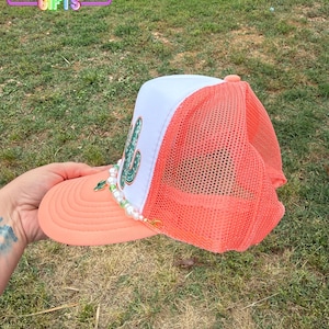 May include: A coral and white trucker hat with a cactus design. The hat has a coral brim and mesh back, a white front panel with a green cactus, and a beaded accent. The text "RAINBOW Emporium GIFTS" is visible.