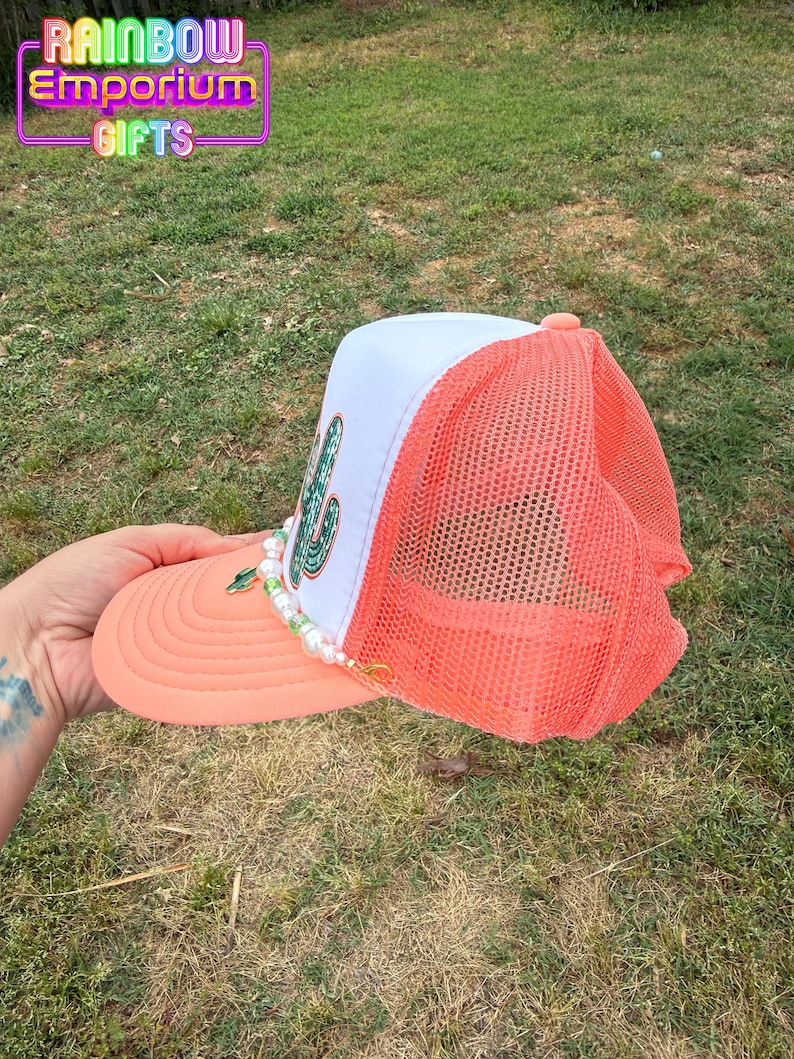 May include: A coral and white trucker hat with a cactus design. The hat has a coral brim and mesh back, a white front panel with a green cactus, and a beaded accent. The text "RAINBOW Emporium GIFTS" is visible.