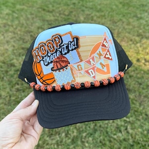 May include: Black and light blue trucker hat with a basketball theme. The hat features the text "HOOP there it is!" and "GAME DAY" on the front, along with basketball graphics and a beaded trim. The hat is a great accessory for basketball fans.