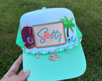 May include: A turquoise and white baseball cap with the word "Salty" embroidered on the front. The cap features a seashell and palm tree design, with a beaded trim and seashell charms. The cap has a mesh back and a curved brim.