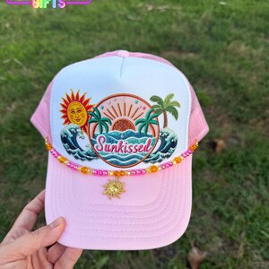 May include: Pink and white trucker hat with a beaded sun charm. The hat features a graphic of a sun, palm trees, and ocean waves with the word "Sunkissed". The top of the hat has a pink mesh backing and the text "Rainbow Emporium Gifts".