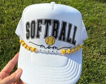 May include: White trucker hat with the word "SOFTBALL" in black block letters. A yellow softball graphic is centered below the word. The word "mom" is below the softball graphic. A beaded necklace with yellow and white beads is attached to the brim.