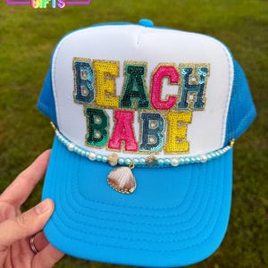 May include: A blue and white trucker hat with the words "BEACH BABE" in colorful sequined letters. The hat features a pearl and seashell embellishment along the brim. The top left corner of the image has the text "RAINBOW EMPORIUM GIFTS".
