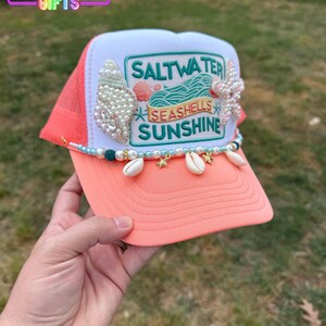 May include: Coral and white trucker hat with the text "Saltwater, Seashells, Sunshine". The hat is decorated with seashells, pearls, and a beaded chain with seashell charms. The hat has a mesh back.