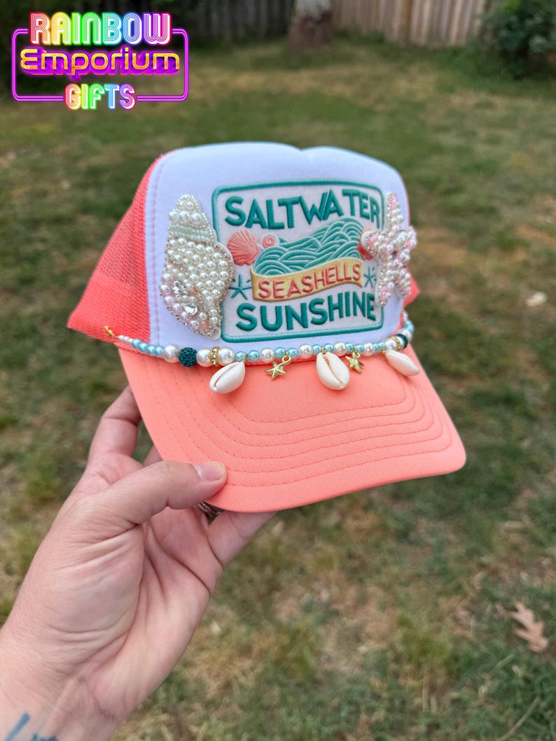 May include: Coral and white trucker hat with the text "Saltwater, Seashells, Sunshine". The hat is decorated with seashells, pearls, and a beaded chain with seashell charms. The hat has a mesh back.