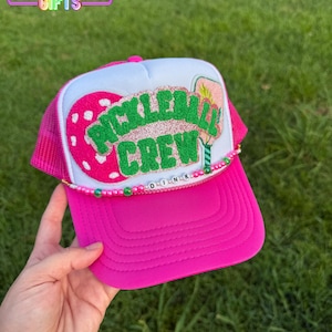 May include: A pink trucker hat with a white mesh back and a curved brim. The hat features the text "PICKLEBALL CREW" in green glitter and a beaded "DINK" accent. The hat is held by a hand against a green grass background.