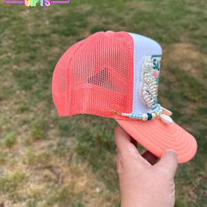 May include: A coral and white trucker hat with a mesh back and a curved brim. The hat features a beaded embellishment and the word "SAL" on the front. The Rainbow Emporium Gifts logo is visible in the top left corner.