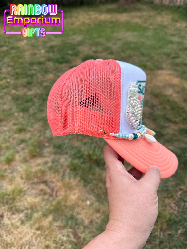 May include: A coral and white trucker hat with a mesh back and a curved brim. The hat features a beaded embellishment and the word "SAL" on the front. The Rainbow Emporium Gifts logo is visible in the top left corner.