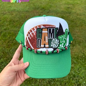 May include: Green and white trucker hat with a football design and the words "GAME DAY." The hat features a beaded trim and a foam finger. The top left corner of the image has the text "RAINBOW EMPORIUM GIFTS."