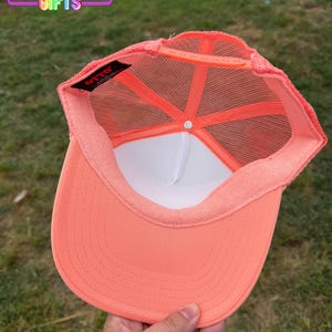 May include: A coral-colored trucker hat with a white interior and mesh backing. The hat features a curved brim and a snap closure. The text "Rainbow Emporium Gifts" is visible in the upper left corner.