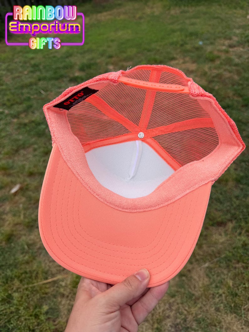 May include: A coral-colored trucker hat with a white interior and mesh backing. The hat features a curved brim and a snap closure. The text "Rainbow Emporium Gifts" is visible in the upper left corner.
