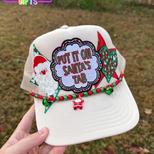 May include: A cream-colored trucker hat with a mesh back and a festive Christmas theme. The hat features a patch with the text "PUT IT ON SANTA'S TAB", along with Santa Claus and decorative elements. A beaded garland with bells and a Santa charm adorns the brim.