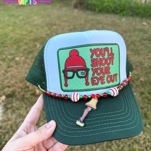 May include: A green and white trucker hat with a patch that reads "You'll Shoot Your Eye Out." The hat features a beaded accent and a charm of a leg lamp. The hat is from Rainbow Emporium Gifts.