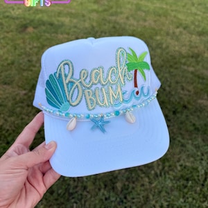 May include: White trucker hat with the text "Beach Bum" in teal and gold embroidery. The hat features a seashell, starfish, and palm tree design, with a beaded accent along the brim. The hat has a white mesh back.