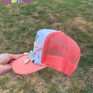 May include: A coral and white trucker hat with a mesh back. The hat features a beaded embellishment with seashells and a jeweled starfish. The text "Rainbow Emporium Gifts" is visible on the top of the hat.