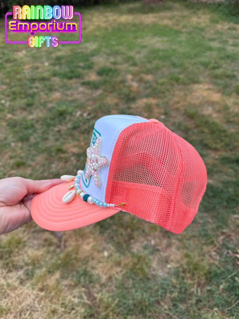 May include: A coral and white trucker hat with a mesh back. The hat features a beaded embellishment with seashells and a jeweled starfish. The text "Rainbow Emporium Gifts" is visible on the top of the hat.
