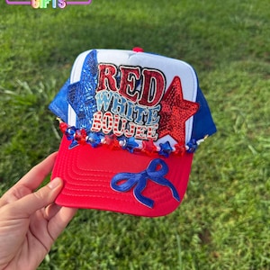 May include: A red, white, and blue baseball cap with the text "RED WHITE BOUIJEE". The cap features a red brim, a white crown, and blue mesh sides. Embellishments include stars and a blue bow.