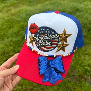 May include: A white, red, and blue trucker hat with a red lip, stars, and the text "American Babe" on the front. The hat has a red bill and a blue bow with sequins.