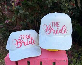 May include: Two white trucker hats with pink glitter lettering. The larger hat says "The Bride" and the smaller hat says "Team Bride".