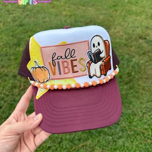 May include: A burgundy and white trucker hat with a fall theme. The hat features a pumpkin and a ghost reading a book, with the words "fall vibes". The brim is burgundy, and the crown is white and burgundy mesh.