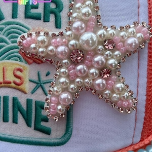 May include: A white and orange trucker hat with a large, embellished starfish. The starfish is adorned with white and pink pearls, pink rhinestones, and a silver-colored trim. The hat also features the text "WATER" and "SHINE".