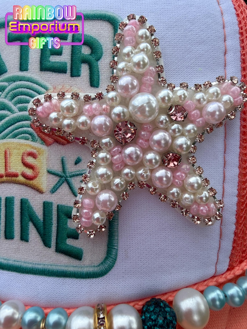 May include: A white and orange trucker hat with a large, embellished starfish. The starfish is adorned with white and pink pearls, pink rhinestones, and a silver-colored trim. The hat also features the text "WATER" and "SHINE".