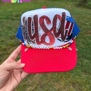 May include: A red, white, and blue trucker hat with the word "USA" in sequined letters. The hat has a red brim, a white front panel, and a blue mesh back. The text "AMERICAN GIRL" is spelled out on the hat.