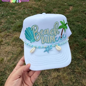 May include: White trucker hat with "Beach Bum" text, a seashell, palm tree, and starfish design. The hat features a beaded accent with seashells and a mesh back. The text "Rainbow Emporium Gifts" is in the upper left corner.