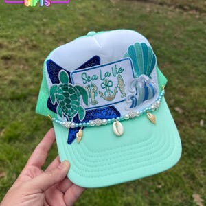 May include: A sea-themed trucker hat with a mint green brim and mesh. The hat features a white front panel with a "Sea La Vie" patch, a turtle, and seashell accents. The hat is decorated with pearls and small seashells.