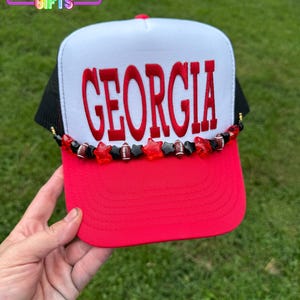 May include: A white and red trucker hat featuring the word GEORGIA in bold red letters across the front panel. The hat is accented with a beaded trim consisting of red star-shaped beads, black star-shaped beads, and small football-shaped beads.