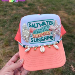 May include: A coral and white trucker hat with the text "Saltwater, Seashells, Sunshine." The hat is decorated with seashells, pearls, and a starfish. The brim is coral, and the mesh backing is also coral.