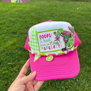 May include: A bright pink and white trucker hat featuring a rectangular patch with the text "OOOPS! I had another MARGARITA" alongside an embroidered margarita glass, a lime slice, and a cocktail umbrella, accented with a beaded trim and lime charm.