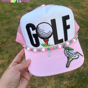 May include: Pink and white trucker hat with the word "GOLF" in large black letters. The hat features a golf ball on a tee, a beaded accent, and a margarita patch. The text "Rainbow Emporium Gifts" is visible.