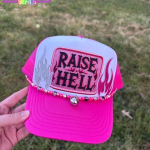 May include: Pink trucker hat with a white front panel featuring a pink and white glitter patch that says "RAISE HELL" with flames on either side. The hat has a silver bell and a beaded trim.